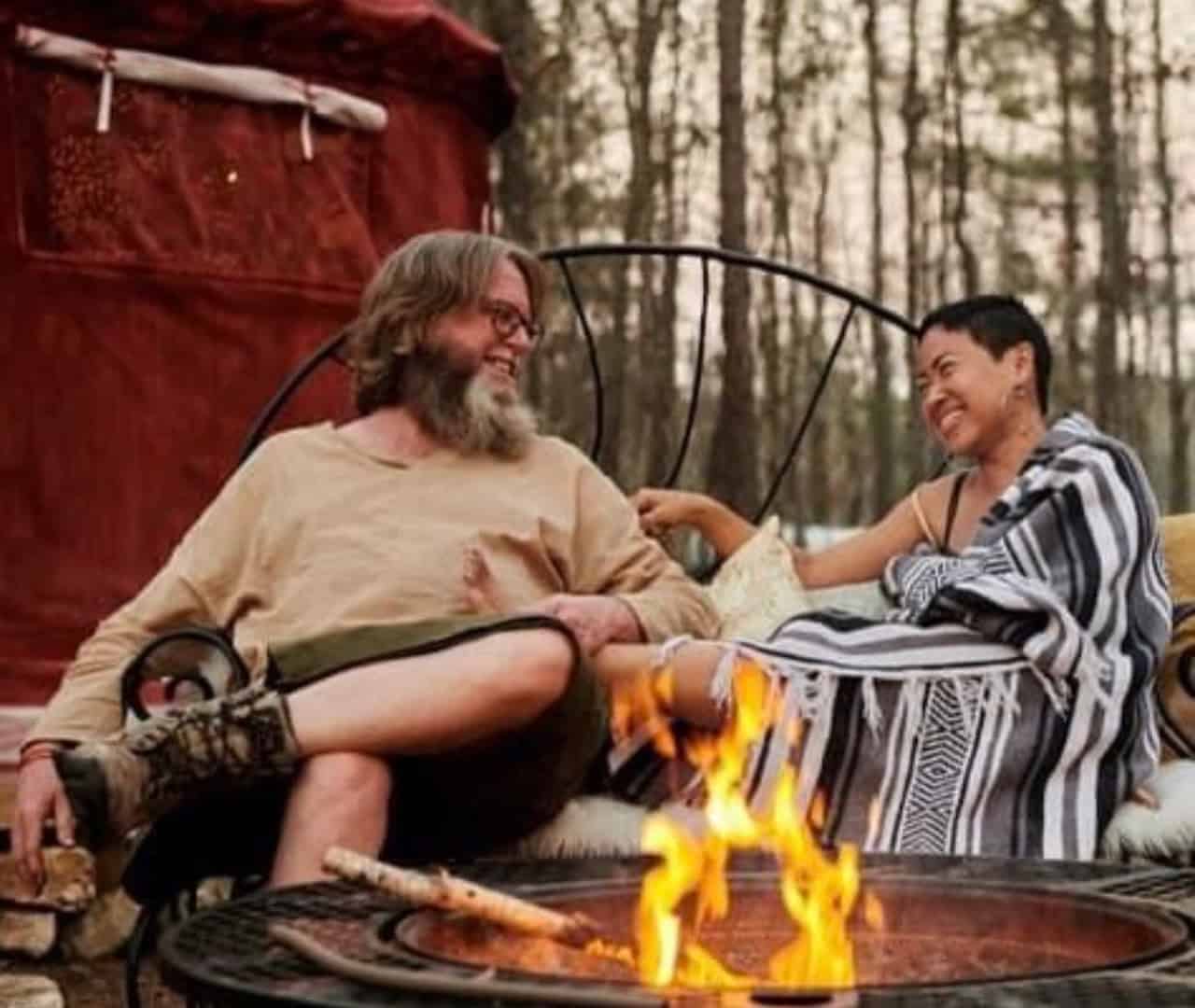 Two people relaxing in chairs beside a firepit outside a red yurt in the woods
