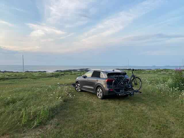 Kia Niro EV stealth camper with bike rack on grassy coastal landscape.
