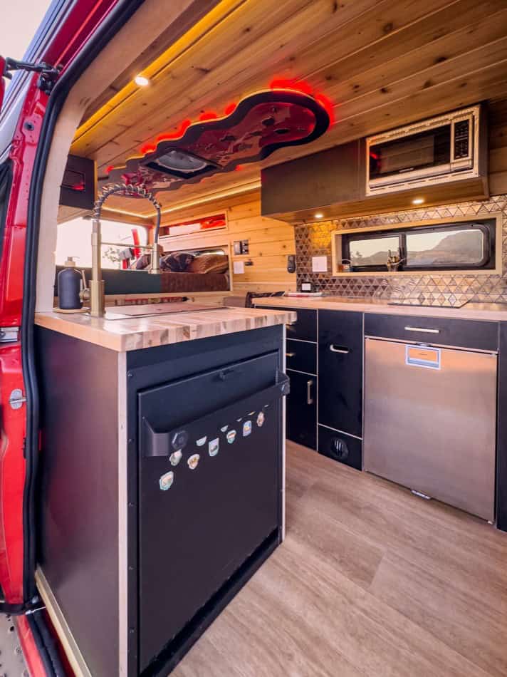 Compact boho-style camper van kitchen with modern appliances and wood accents.