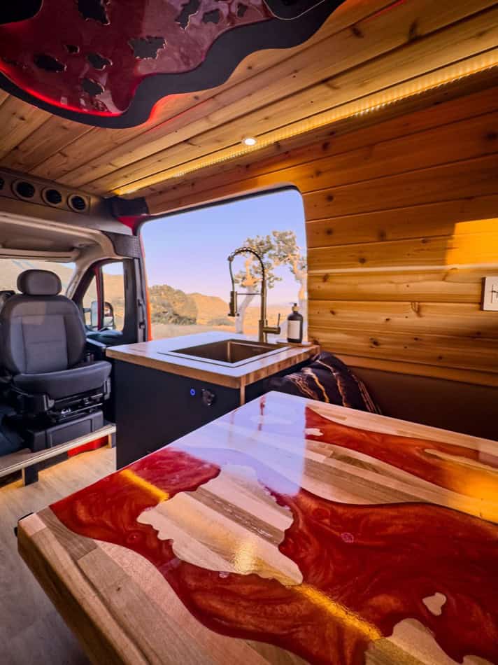 Interior of a boho-style camper van with wooden walls, a small kitchen sink, and scenic outdoor view.