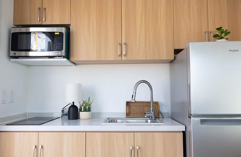 BoxHouse kitchen with light wood cabinets, white countertop, two-burner cooktop, microwave, and stainless steel refrigerator