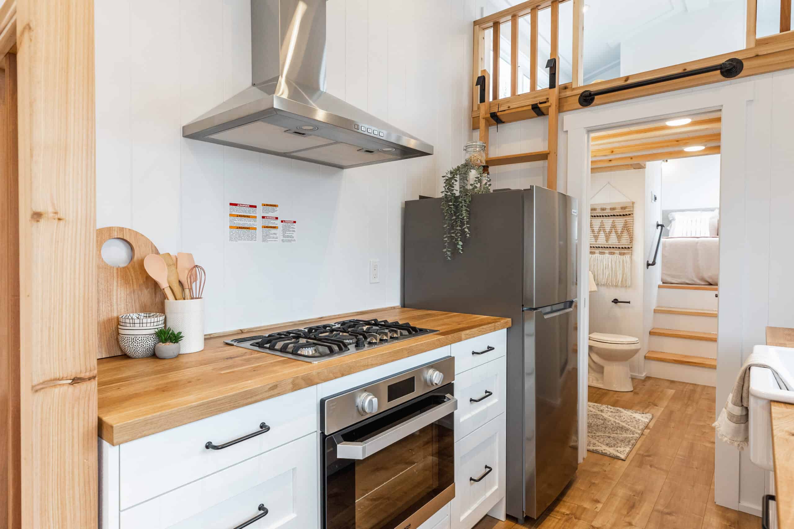 Compact tiny house kitchen with stainless steel stove, refrigerator, and wooden countertops. Bright,.