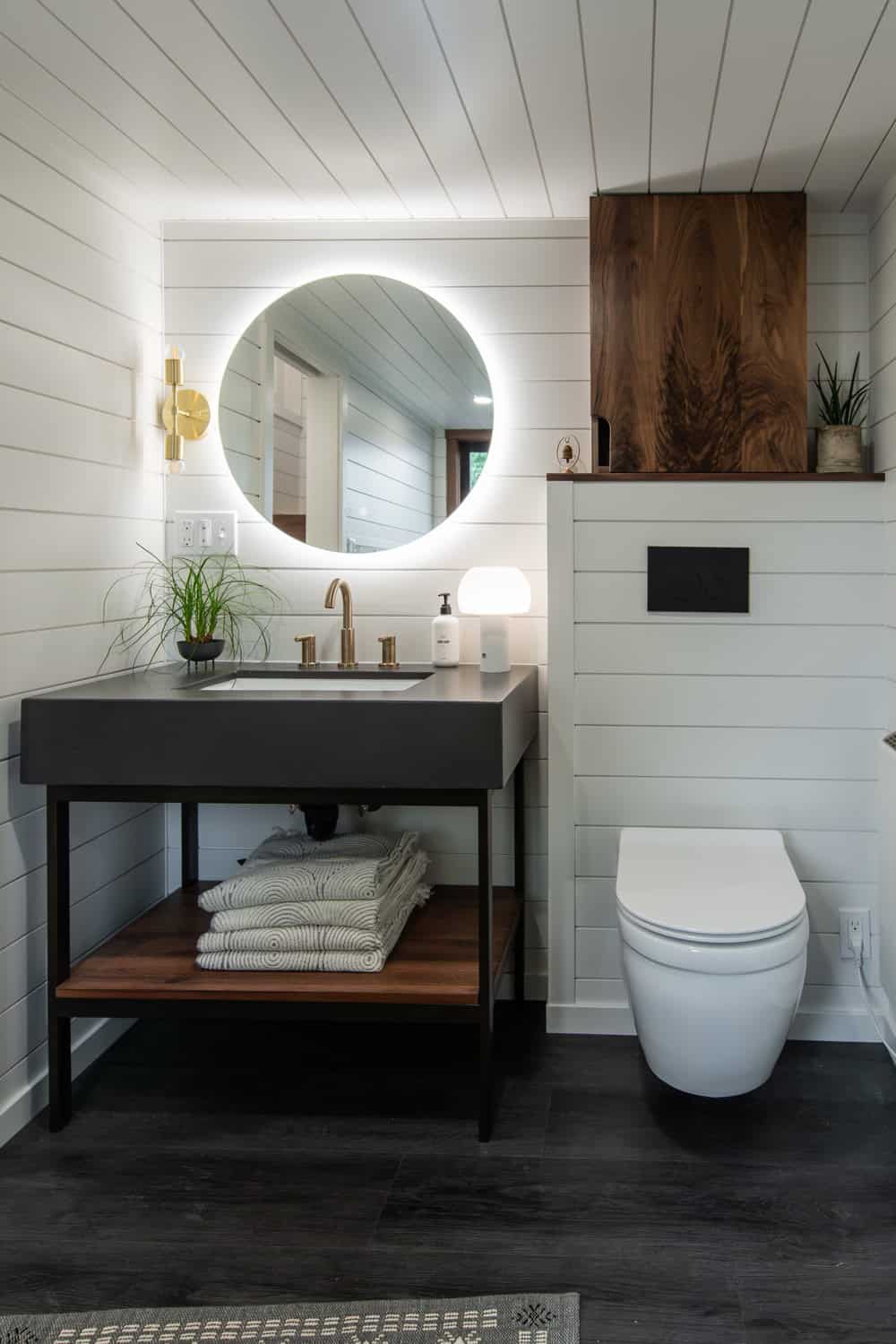 Cascade Max bathroom vanity with a round backlit mirror, black trough sink, walnut medicine cabinet, and wall-hung toilet