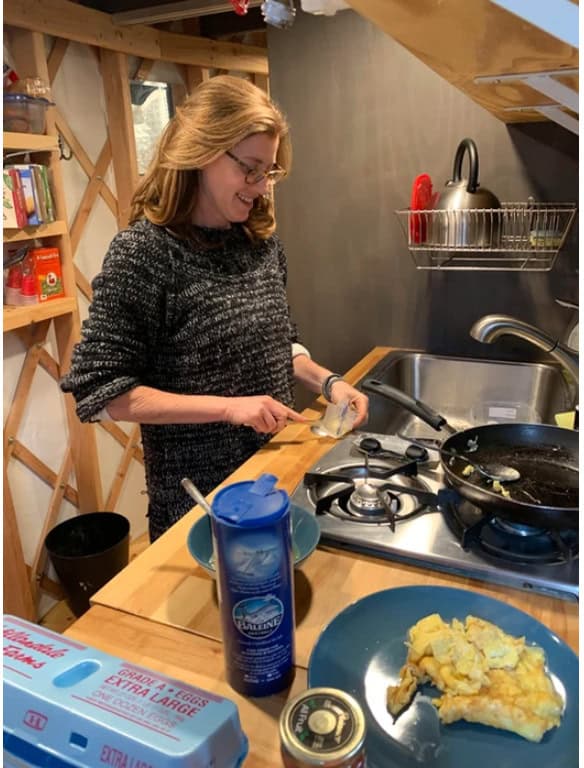 Woman cooking breakfast in a tiny house kitchen with compact appliances and wooden shelves.