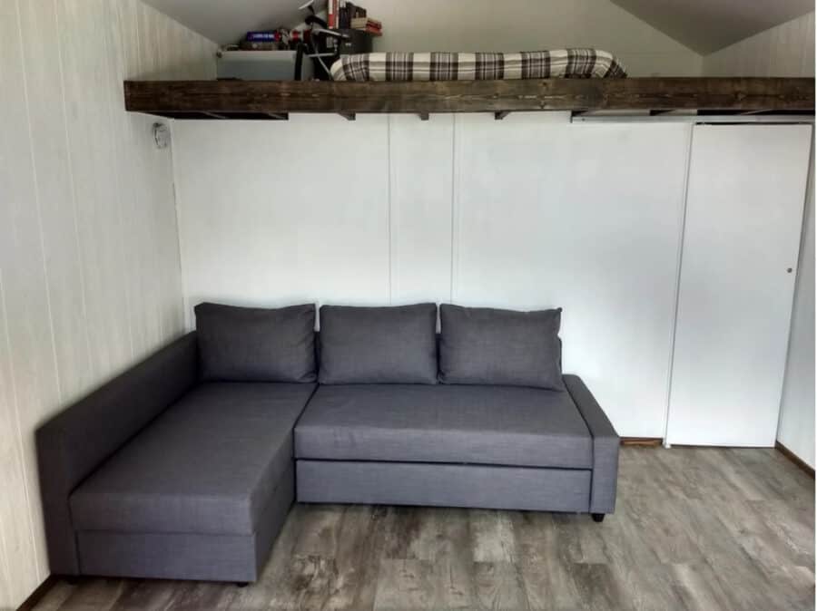 Compact tiny house living space with a gray sectional sofa and loft bed.