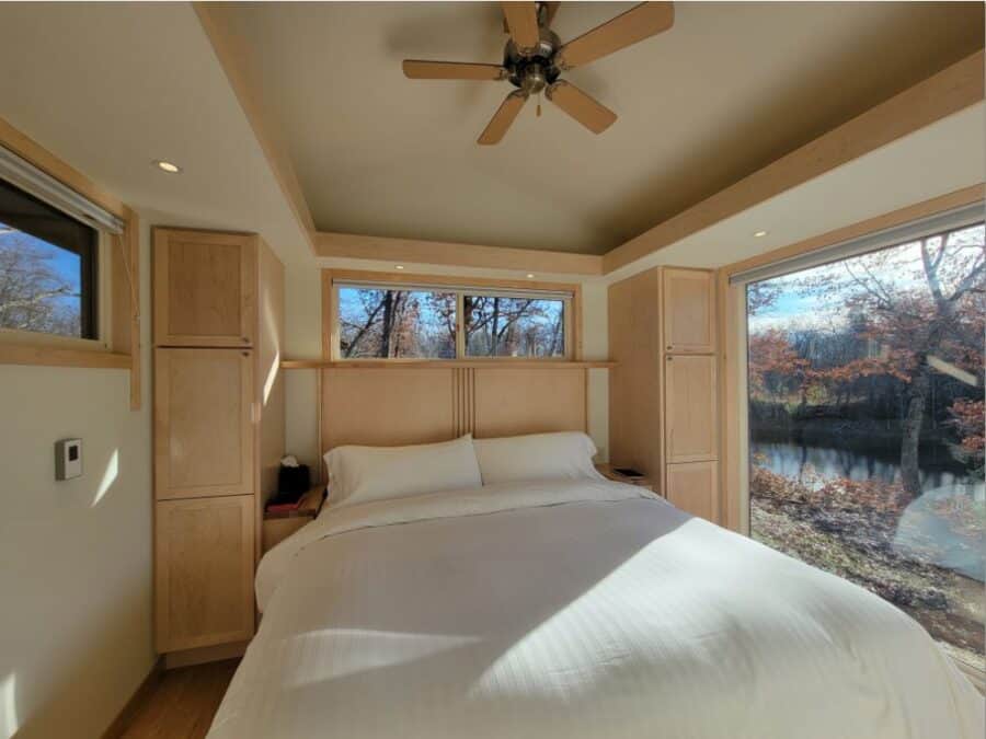 Compact tiny house bedroom with large windows overlooking a serene lake scene.