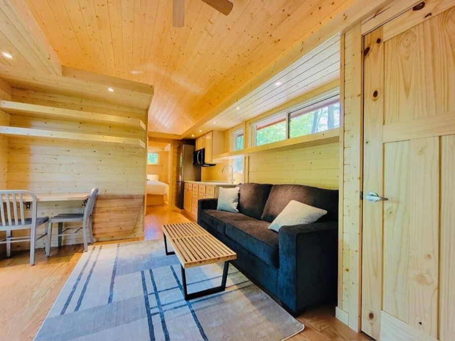 Interior view of a cozy tiny house with wooden walls, a black sofa, and natural light from windows.