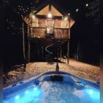 Treehouse with hot tub and cozy lighting in a forest setting.