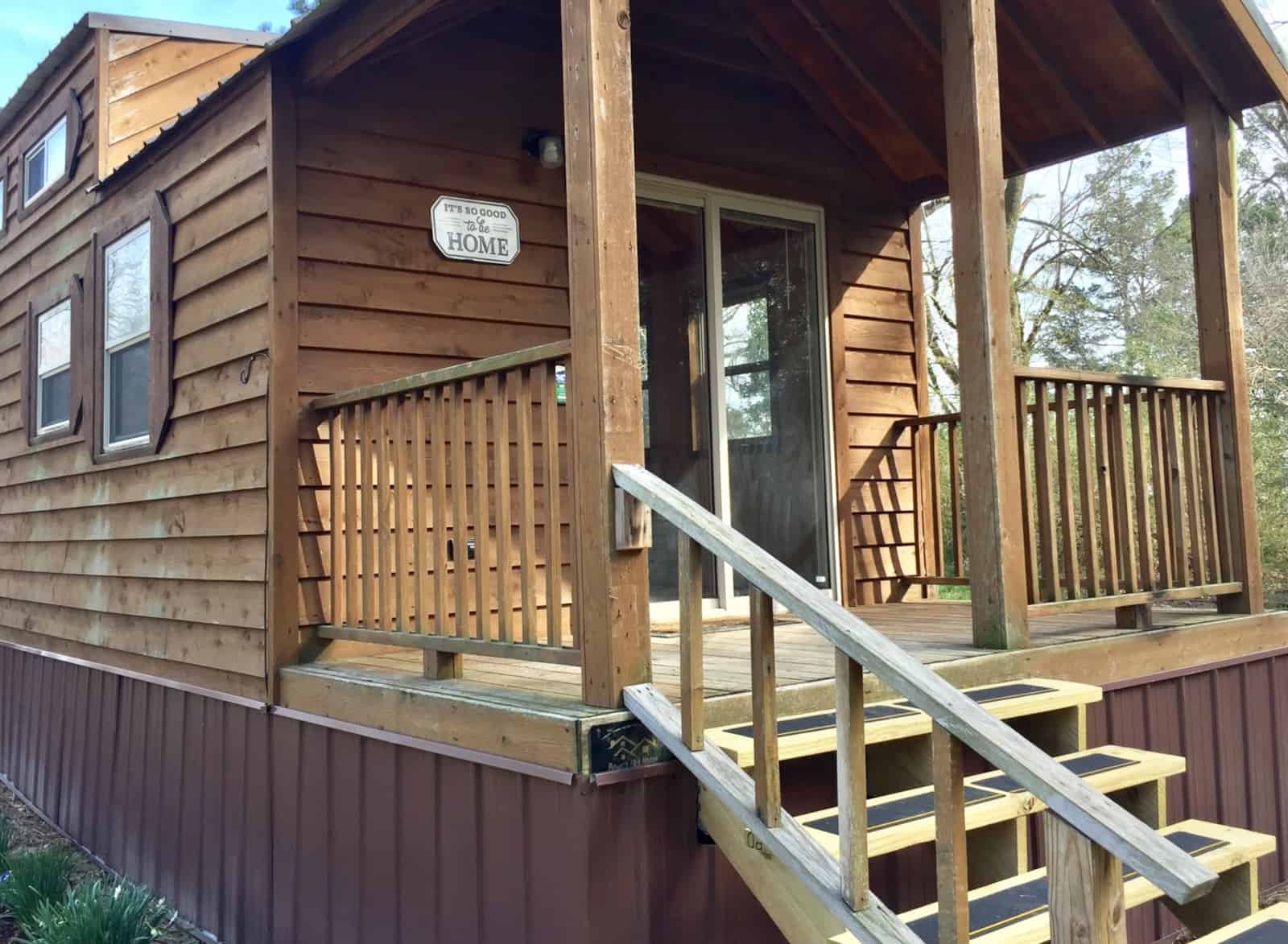 Covered front porch of the Country Cottage tiny home with wood railings, steps, and a sliding glass entry door.