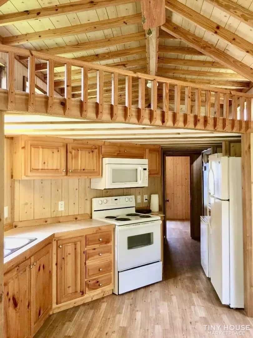 Knotty pine kitchen in the Country Cottage tiny home with a full-size range, microwave, refrigerator, and the loft railing above.