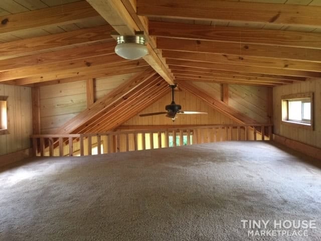 Carpeted upstairs loft with vaulted pine ceiling, ceiling fan, gable windows, and a wood railing overlooking the living area.