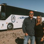 Couple standing outside their tiny house bus in a desert landscape, ready for winter escape.