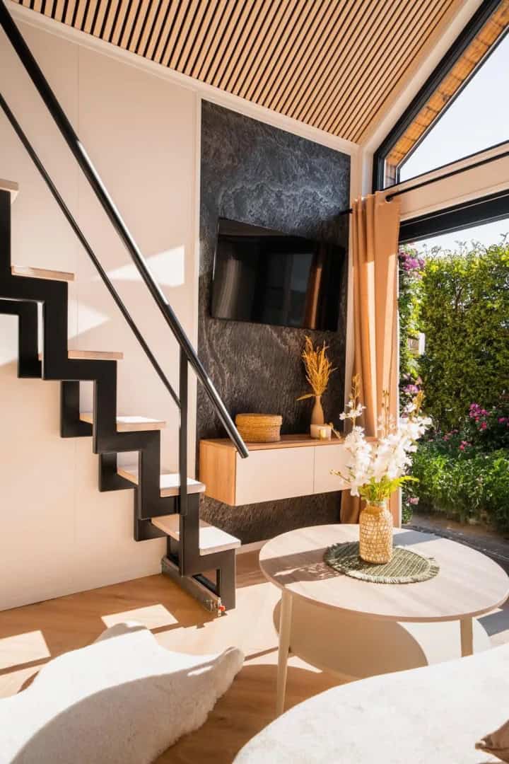 Open-plan living area of the Lukas with a black floating staircase, TV on a stone feature wall, and a dining table