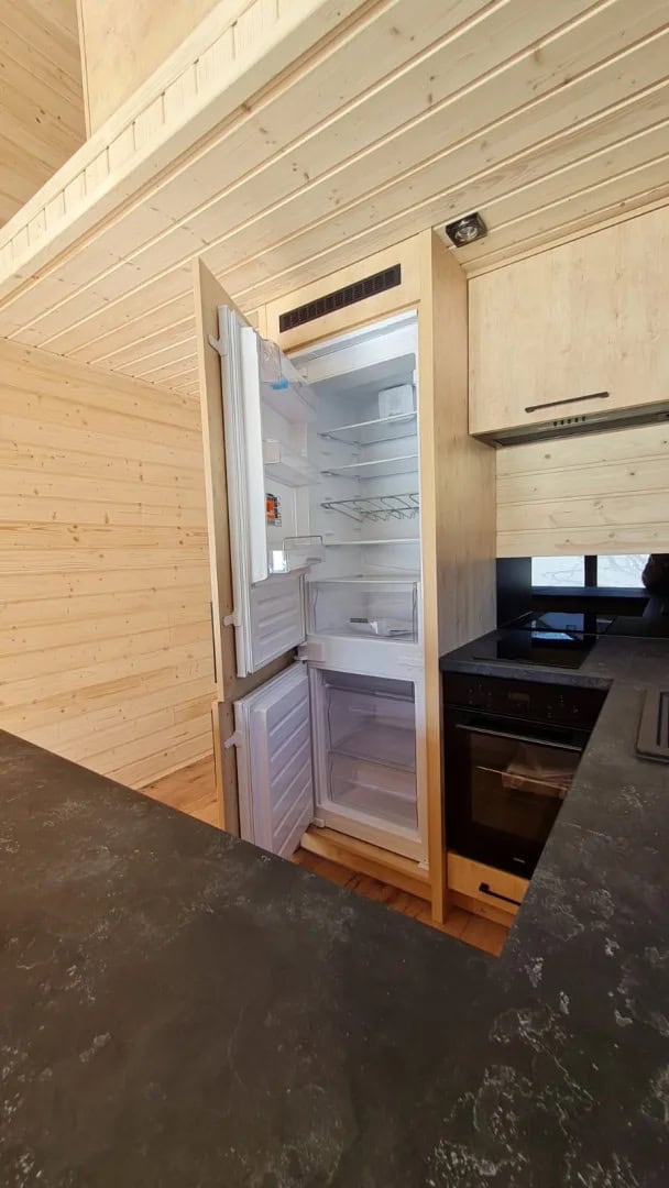 Compact kitchen in the Samuel modular house with a built-in refrigerator and black countertop against spruce walls