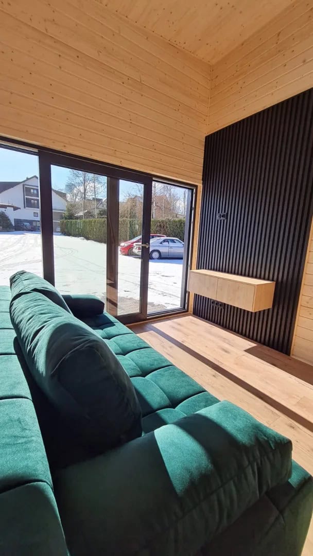 Wood-lined living room of the Samuel modular house with a green sofa, black slat feature wall, and glass sliding doors