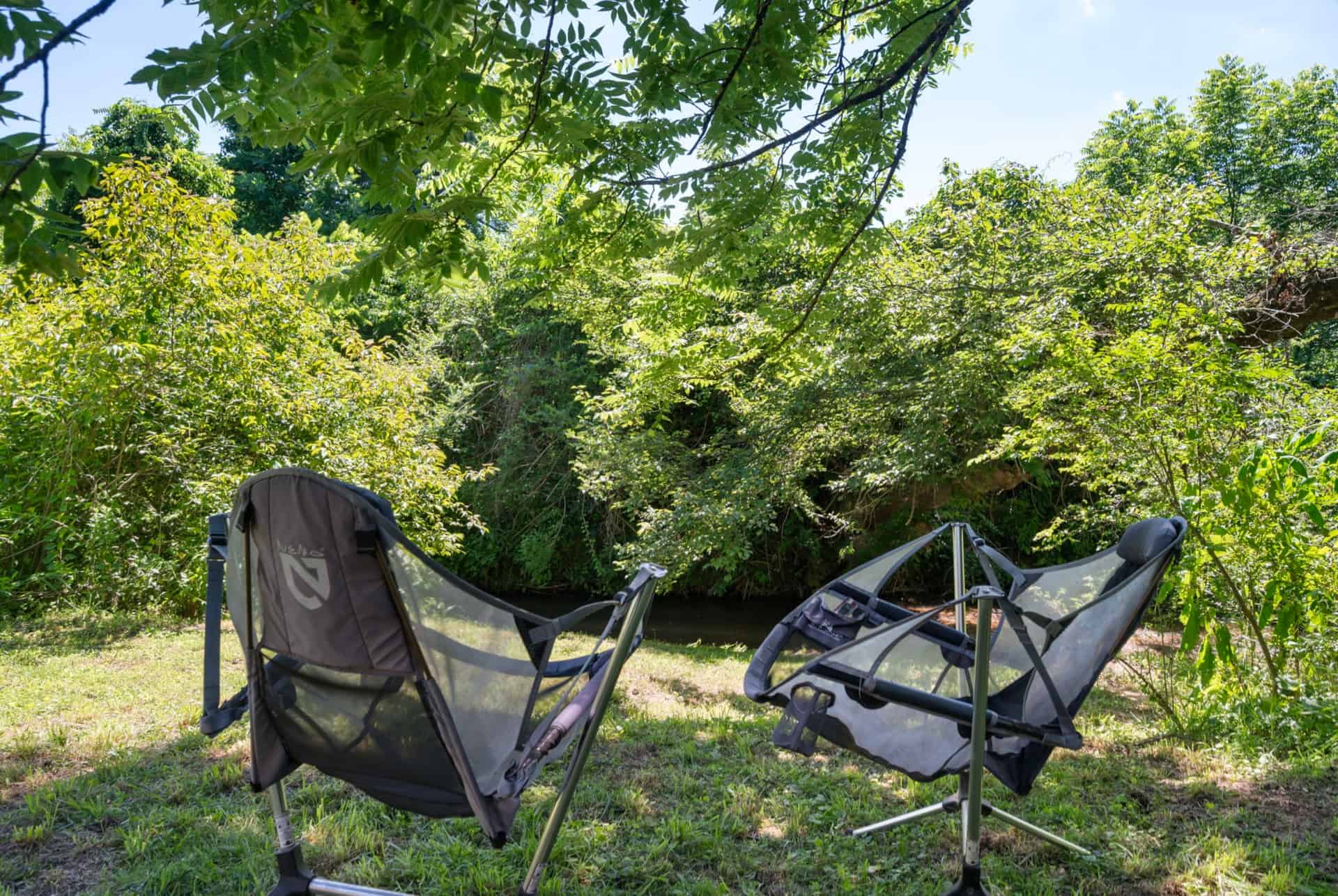 Two reclining camp chairs set up beside the creek under shade trees