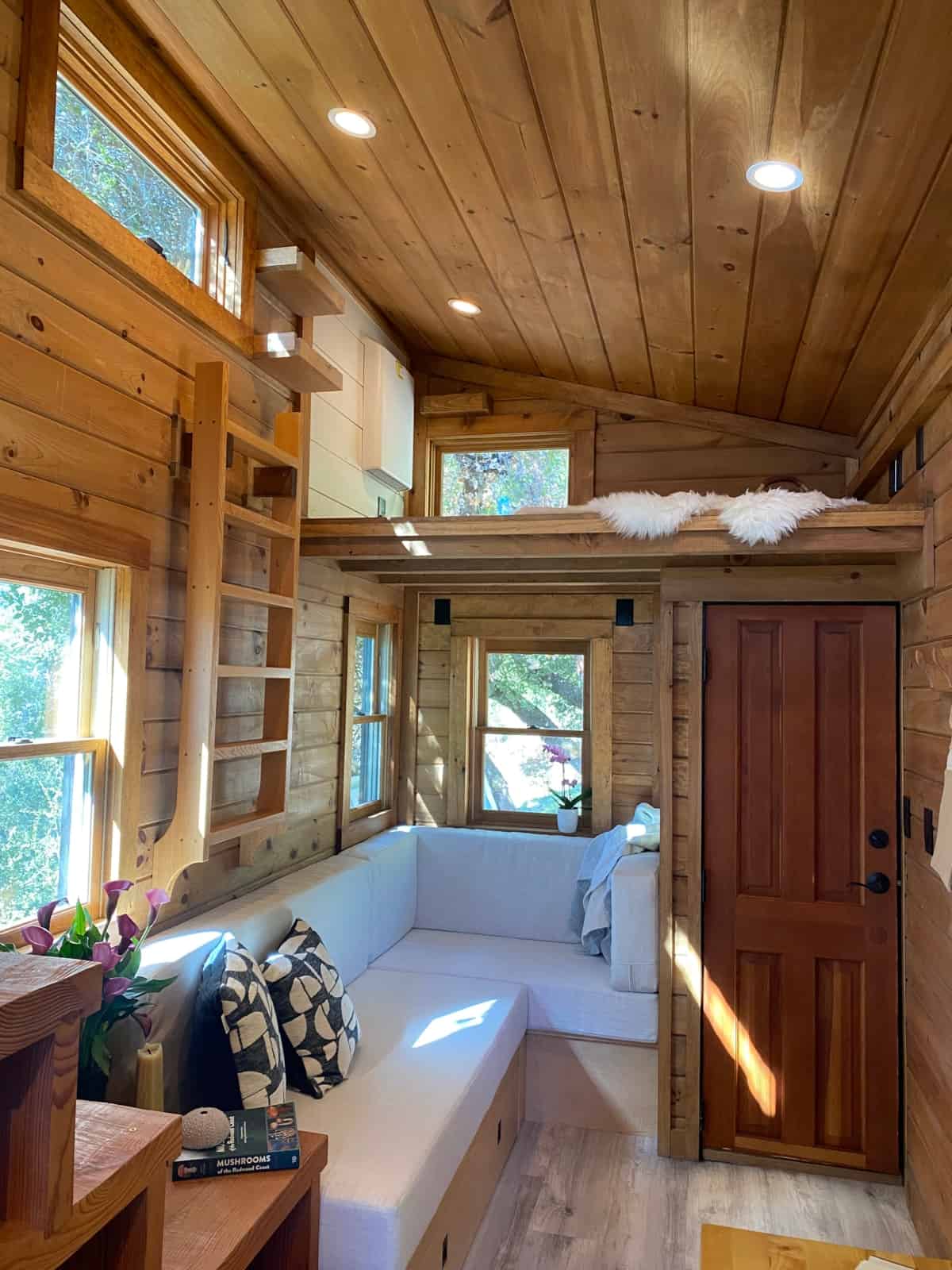 Living room with custom L-shaped sofa, storage drawers, ladder to loft, and windows on three sides