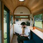 Man working on a laptop inside a tiny house with wooden interior and large windows.