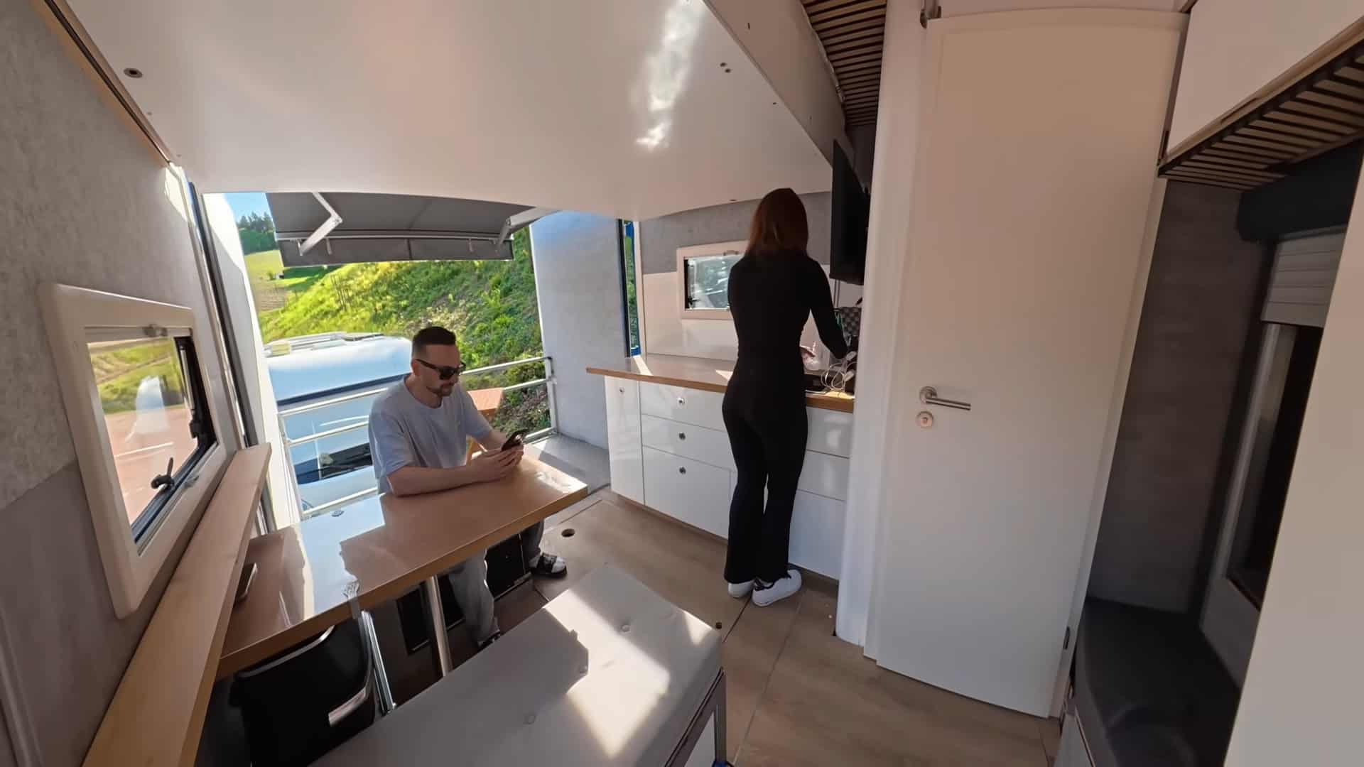 Couple cooking and dining in the semi-truck tiny house with fold-down balcony open and views
