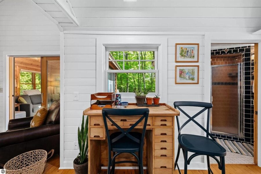 Interior view of a small log cabin with a wooden desk, chairs, and window overlooking greenery.