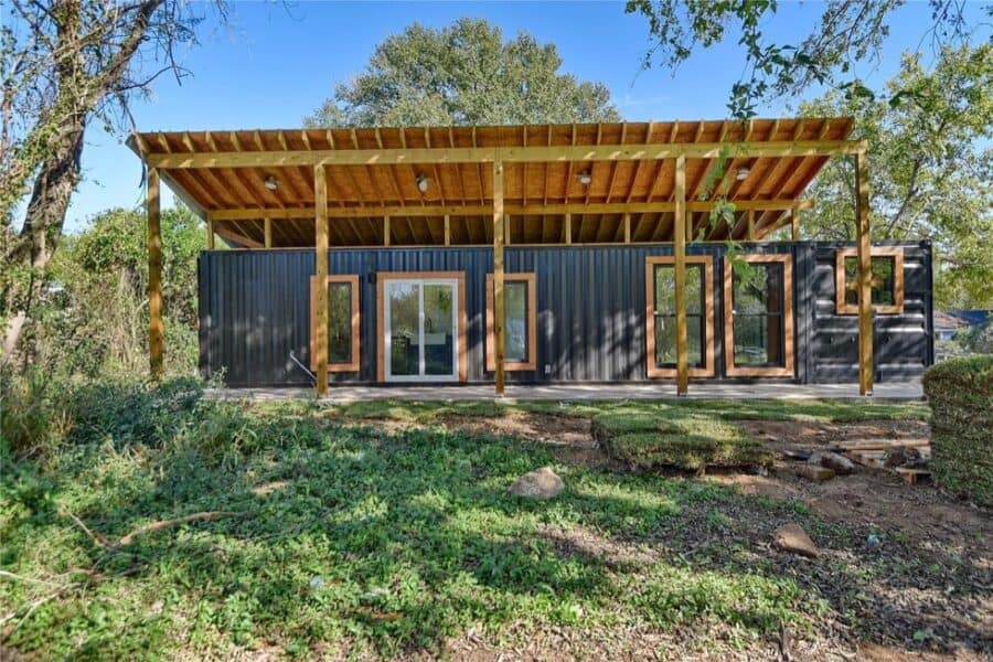 Modern shipping container home with large windows and covered porch in a natural setting.