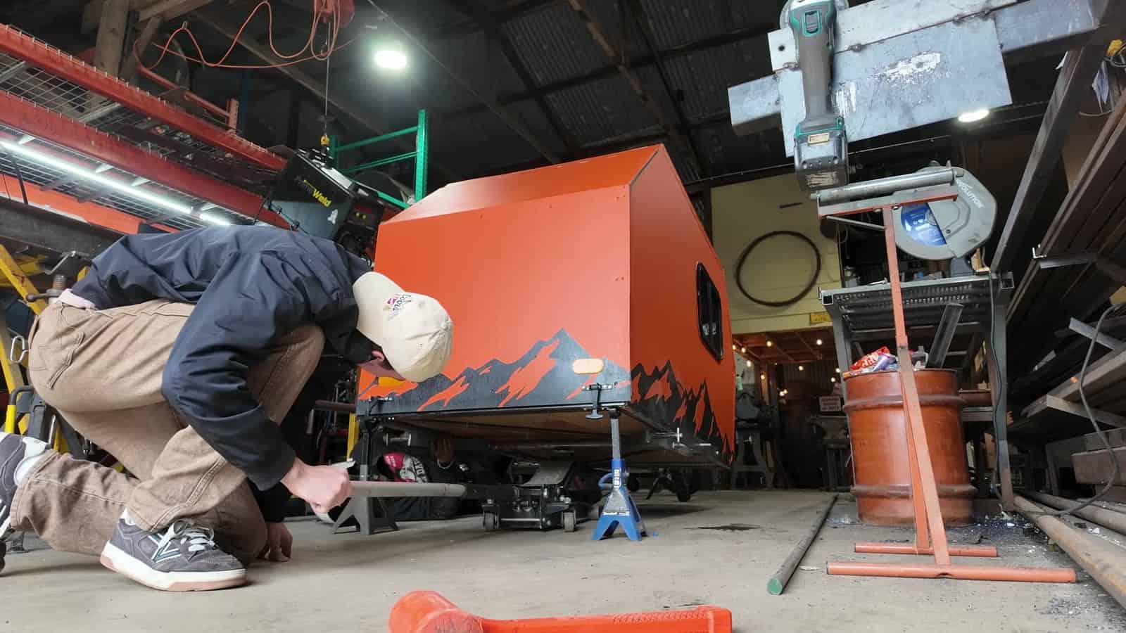 Welder installing a heavier axle and wheel wells under the orange camper in a workshop