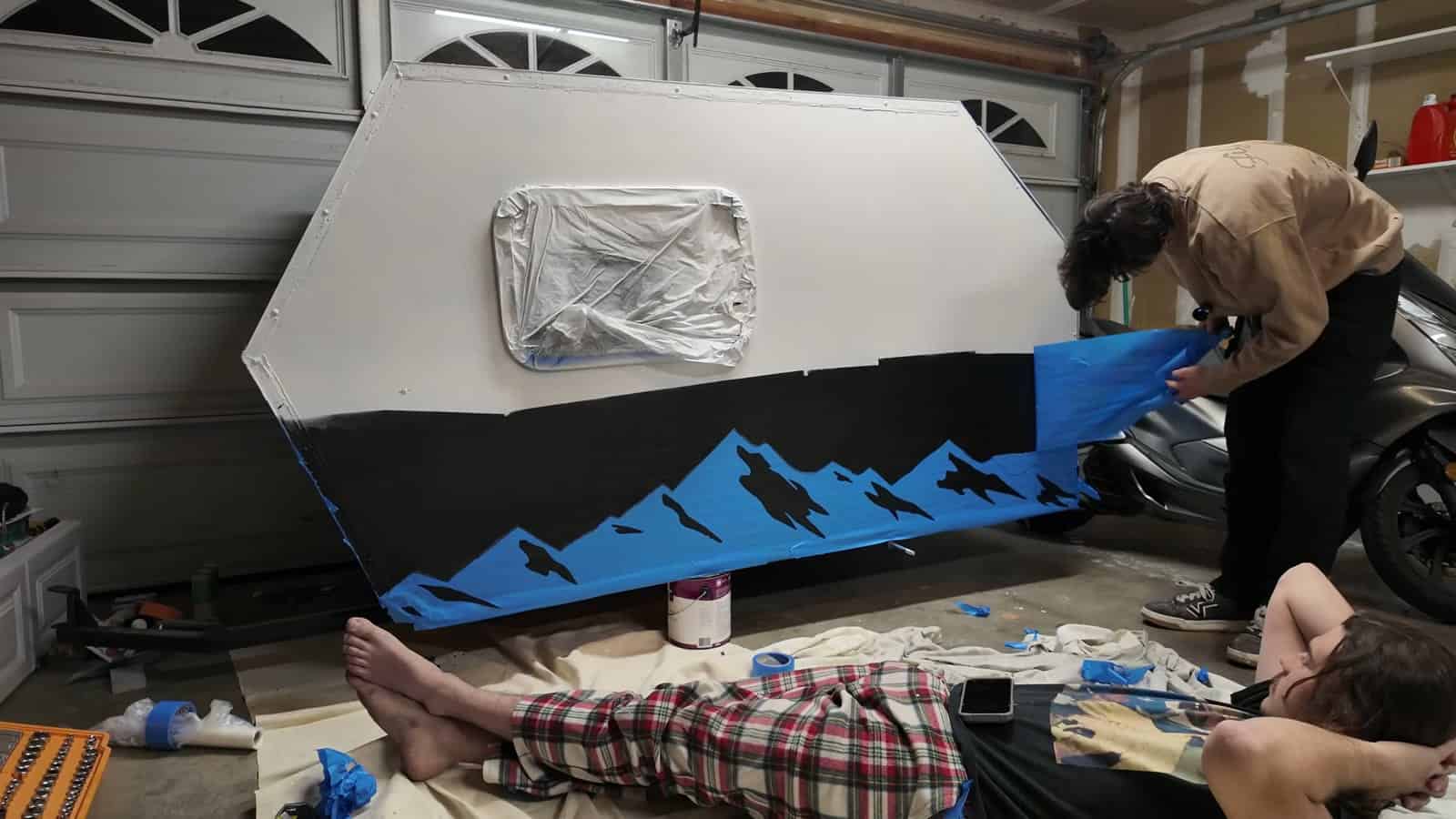 Masking tape mountain stencil being applied along the lower half of the camper in a garage