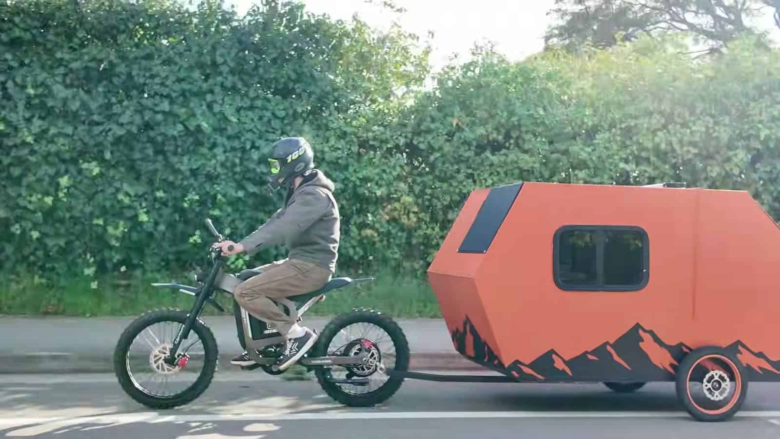 Rider on a Coswheel electric dirt bike towing the orange mountain-graphic camper past a hedge