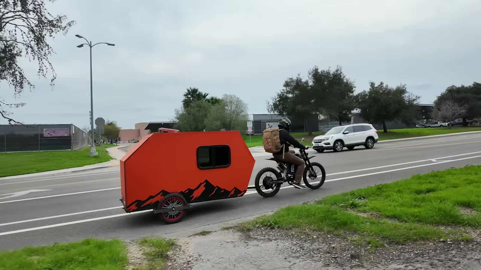 Orange e-bike camper with black mountain graphics being towed down a road by an electric dirt bike