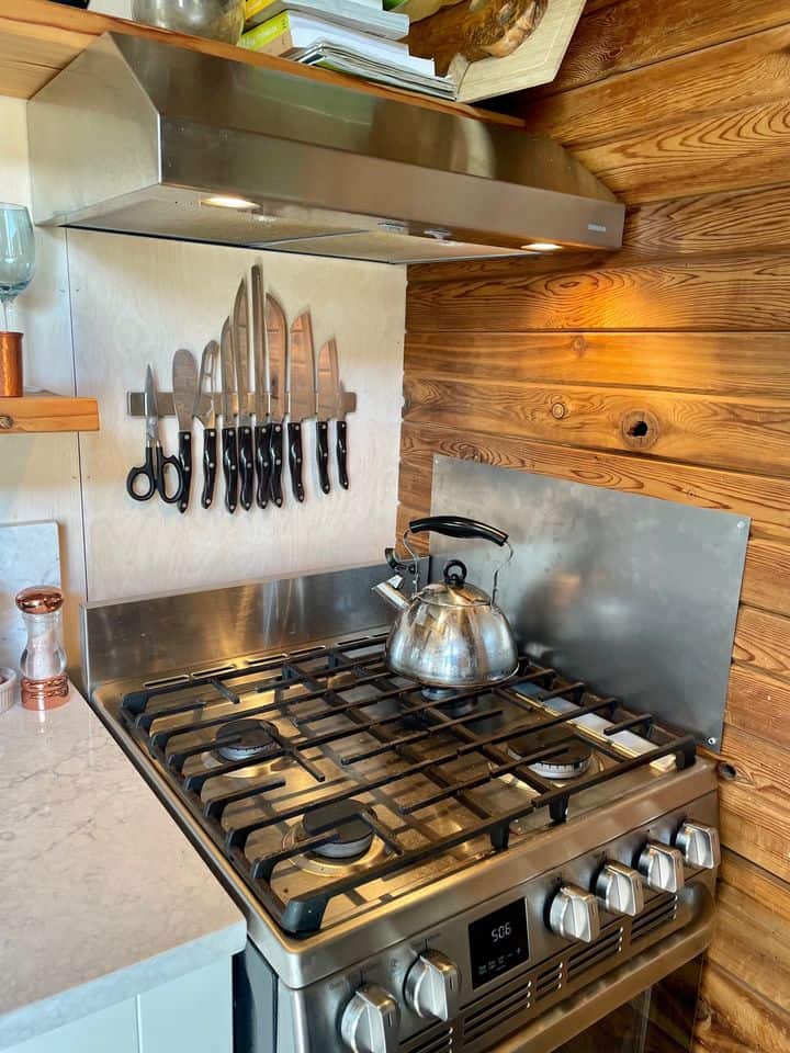 Handcrafted eco-bohemian tiny house kitchen with a wooden wall and a stainless steel stove.