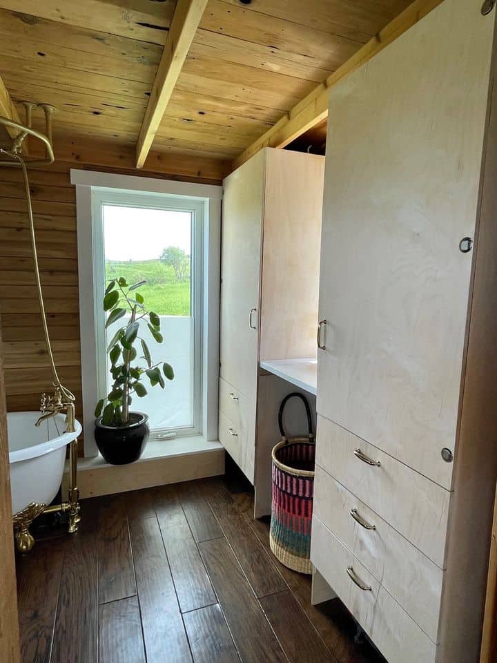 Bright tiny house bathroom with wooden walls, a window overlooking greenery, and minimalist storage.