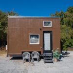 Compact tiny house with a brown exterior, featuring two windows and a white door, set against a back.