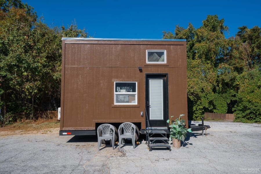 Compact tiny house with a brown exterior, featuring two windows and a white door, set against a back.