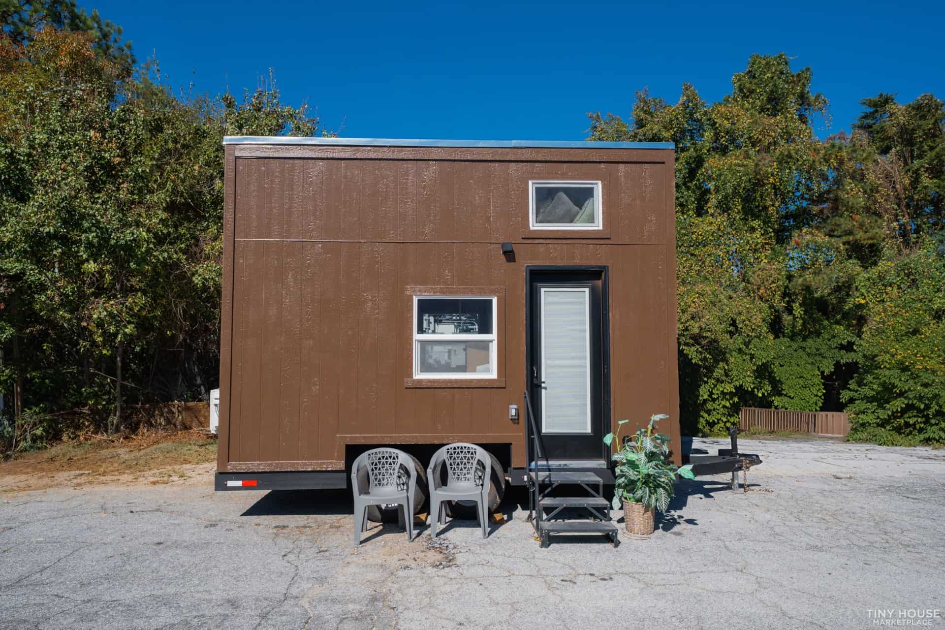 Compact tiny house with a brown exterior, featuring two windows and a white door, set against a back.