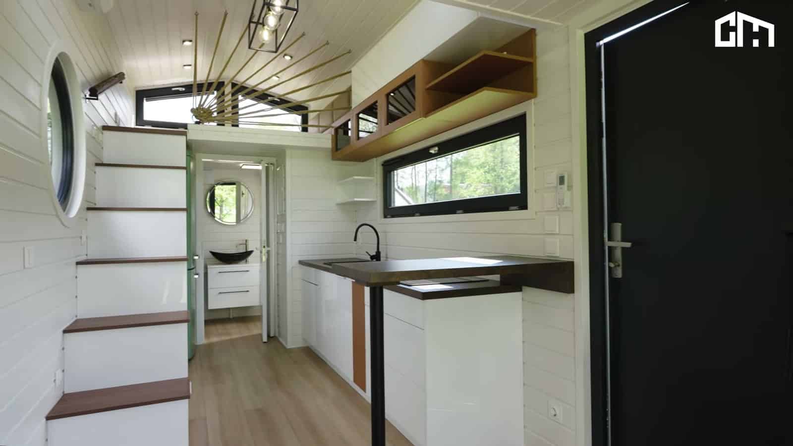 Bright white shiplap interior of the Erica seen from the entry, with storage stairs, a porthole window, the kitchen, and the bathroom at the far end