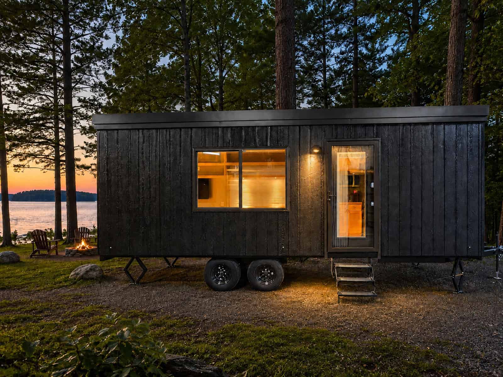Escape eVista tiny house with black charred wood siding parked in the pines beside a lake at sunset