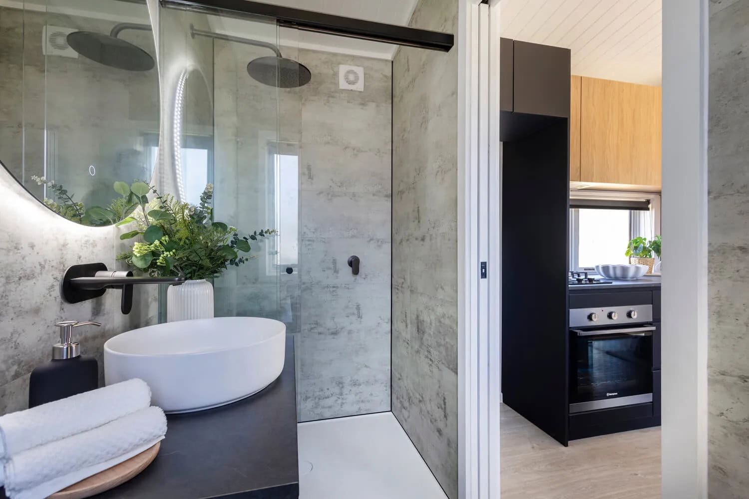 Bathroom in the Esther tiny house with concrete-look wall panels, a rainfall shower behind a frameless glass screen, white vessel basin on a dark benchtop, backlit round mirror, and matte black tapware