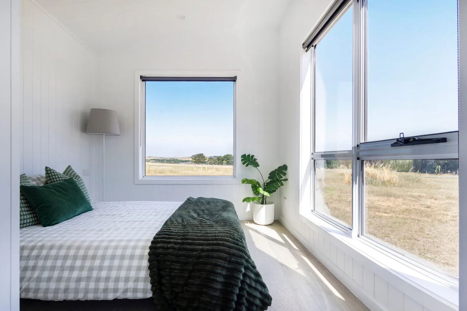 Bedroom of the Esther tiny house with large corner windows framing rural views, white VJ paneling, and timber-look flooring
