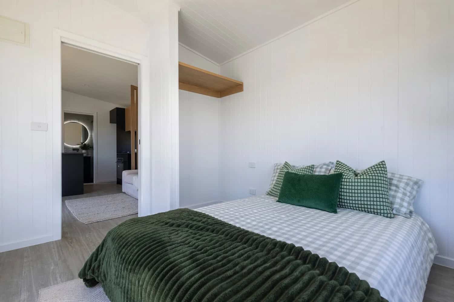 Bedroom in the Esther tiny house with full standing headroom, a floating timber shelf, open wardrobe nook, white VJ paneling, and a doorway through to the living area