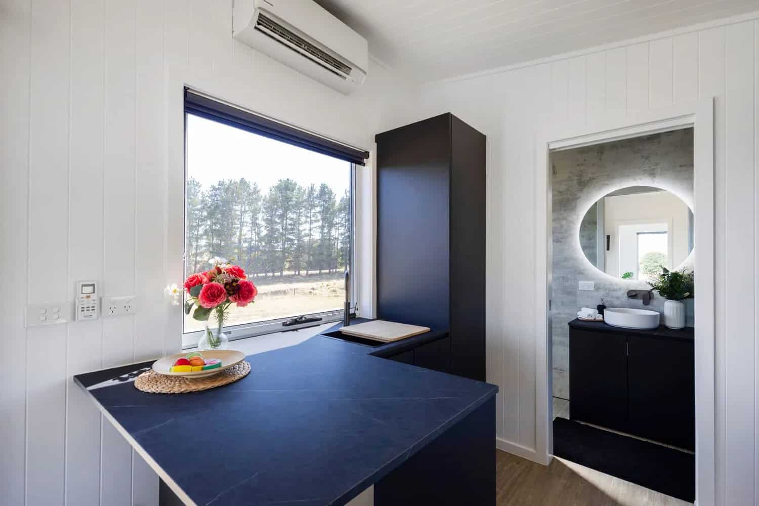 Kitchen peninsula in the Esther tiny house with sink and timber chopping insert, breakfast bar seating, tall black pantry cabinet, and the bathroom doorway with a backlit round mirror beyond