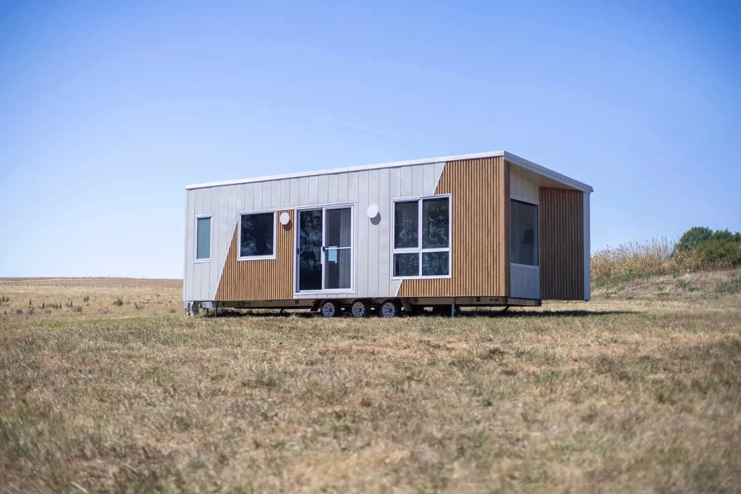 Three-quarter view of the Esther tiny house showing the white standing seam cladding, timber-look batten end wall, and triple-axle trailer with drop-down stabilizing legs