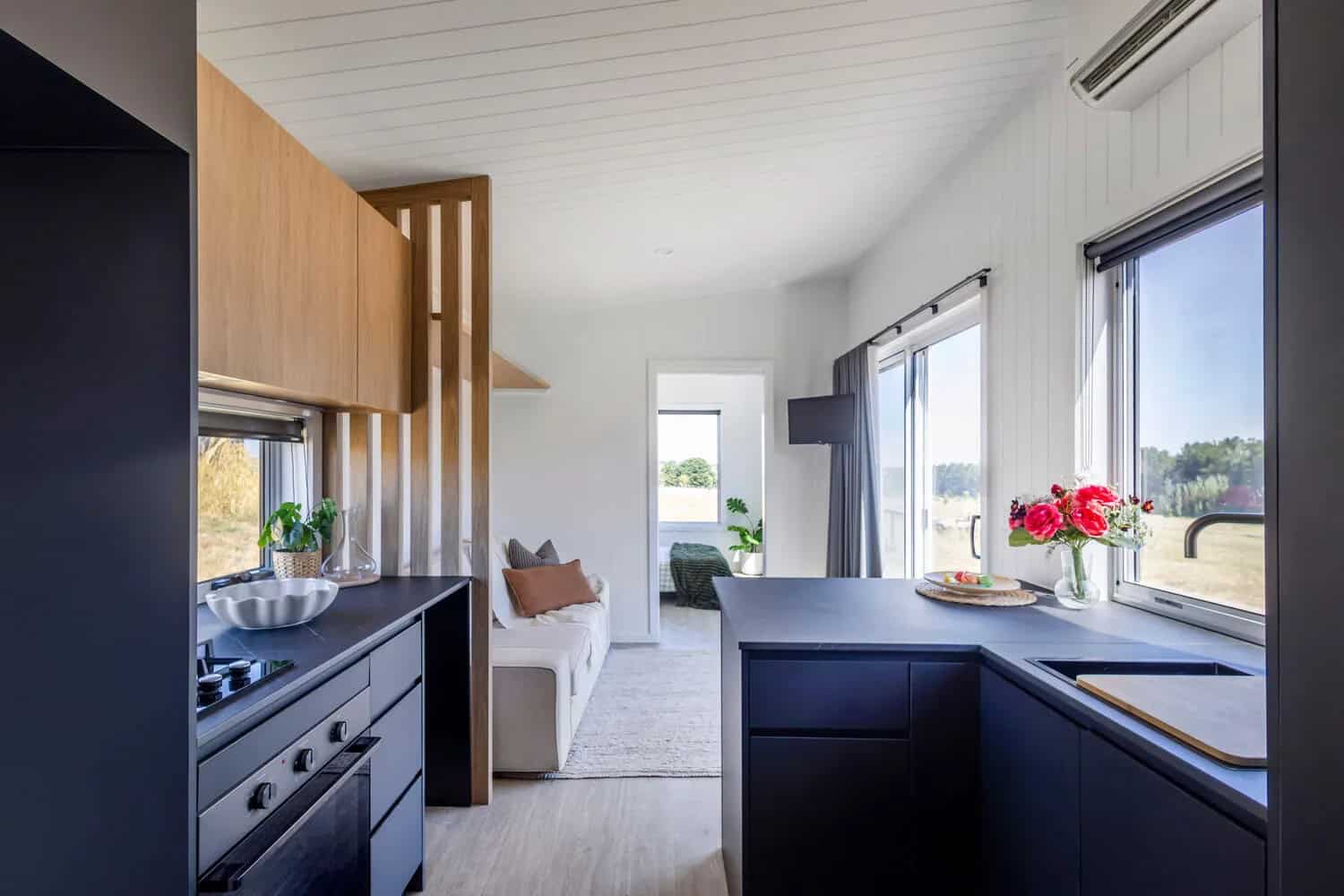 Interior of the Esther tiny house looking from the kitchen through the living area to the bedroom doorway, with black cabinetry, timber upper cupboards, white VJ paneling, and a timber slat screen