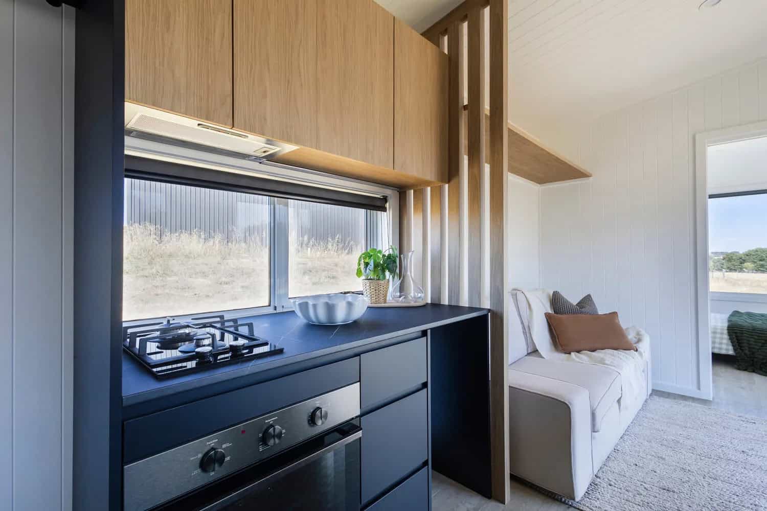 Kitchen in the Esther tiny house with a gas cooktop and under-bench oven, dark stone-look benchtop, pale timber overhead cabinets, concealed rangehood, and a window above the bench