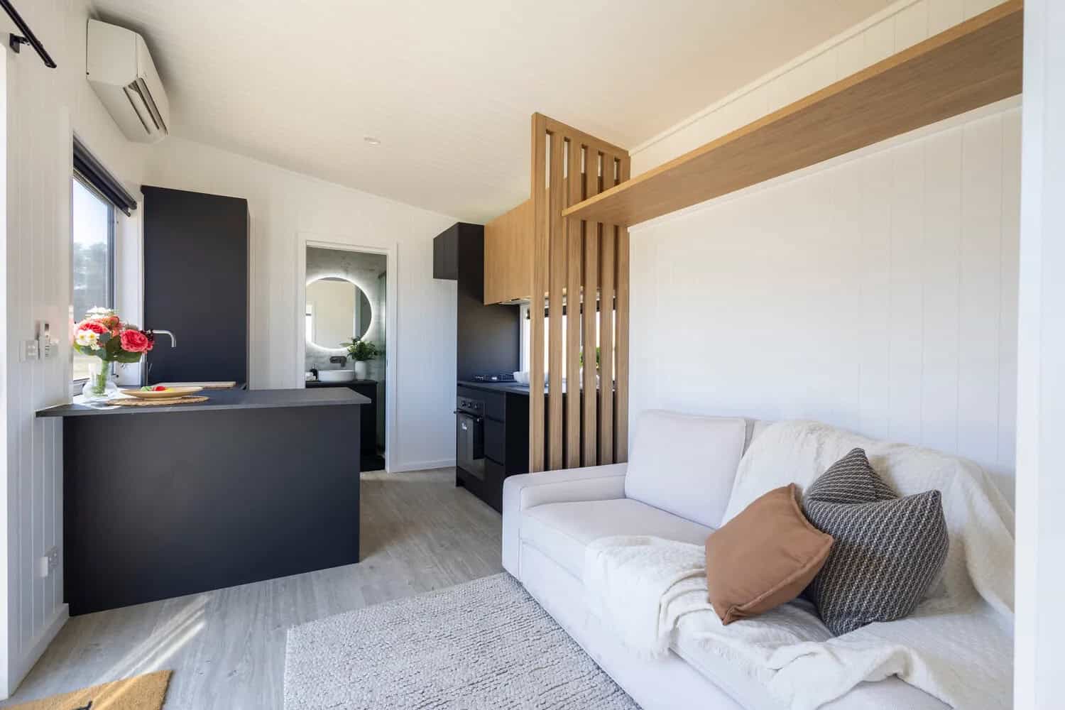 Living area of the Esther tiny house with a white sofa, slatted timber screen and floating shelf, black kitchen peninsula, split-system air conditioner, and the bathroom doorway beyond