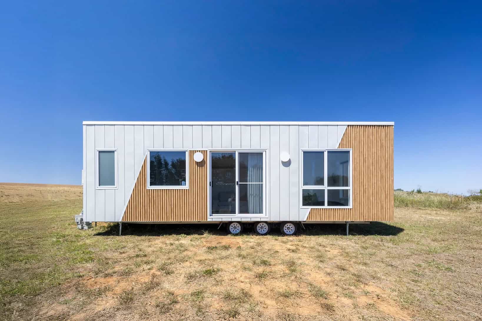 Esther tiny house by CozyCo with white standing seam cladding and timber-look batten panels, sliding glass door and large windows, on a triple-axle trailer in an Australian paddock