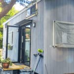 Small blue tiny house with outdoor plants and large window in Australia.