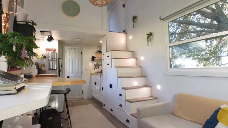 Bright interior of a tiny house featuring built-in stairs, a comfortable sofa, and large windows wit.