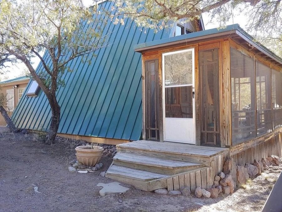 Small A-frame cabin with wooden porch and screened-in area in Texas countryside.