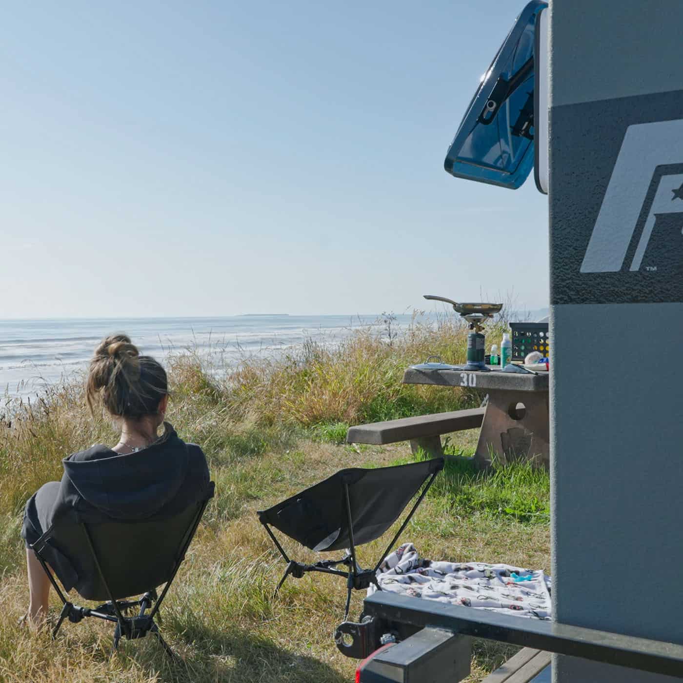 Woman in a camp chair beside a Freedom Camper trailer overlooking the ocean