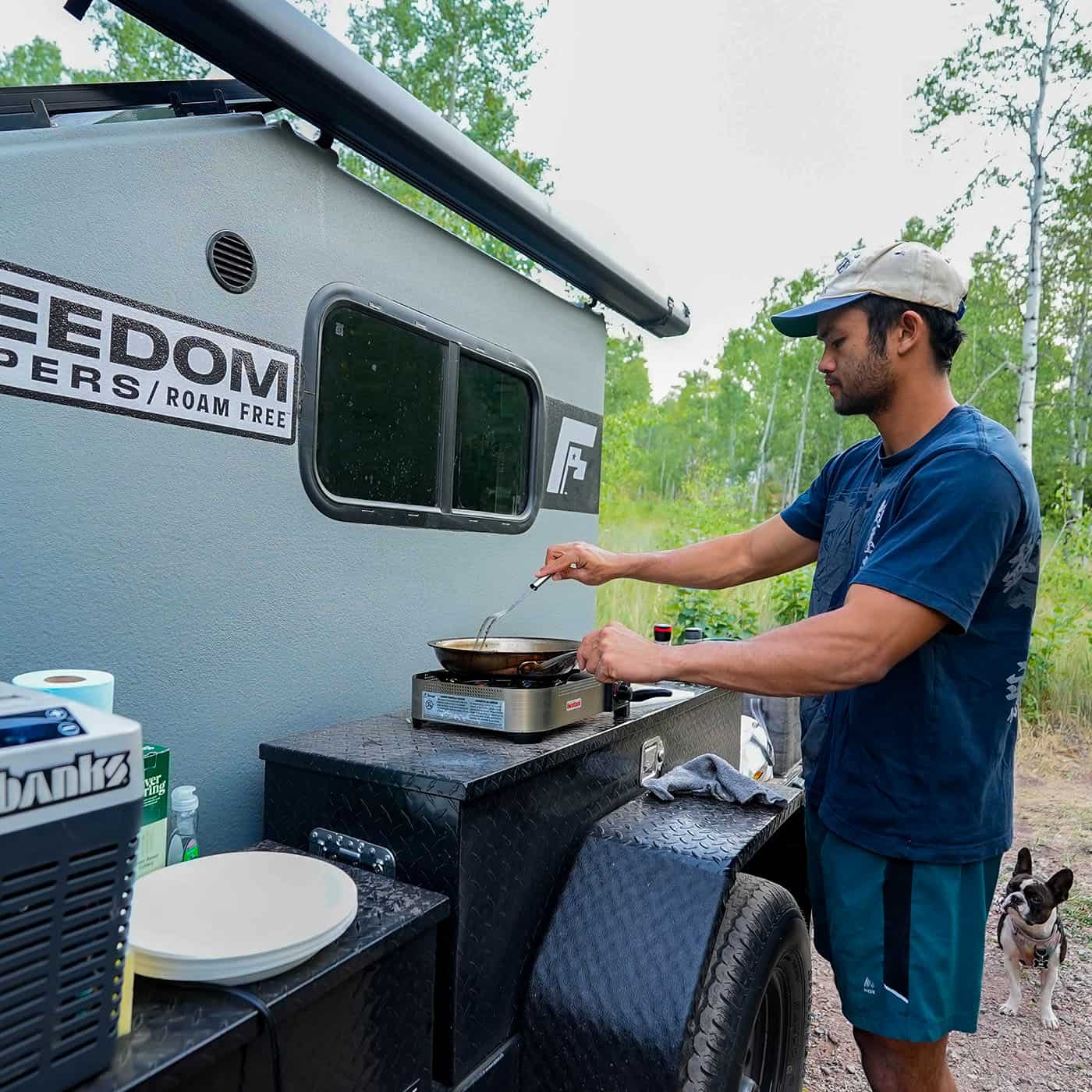 Man cooking on a camp stove beside a Freedom Camper trailer